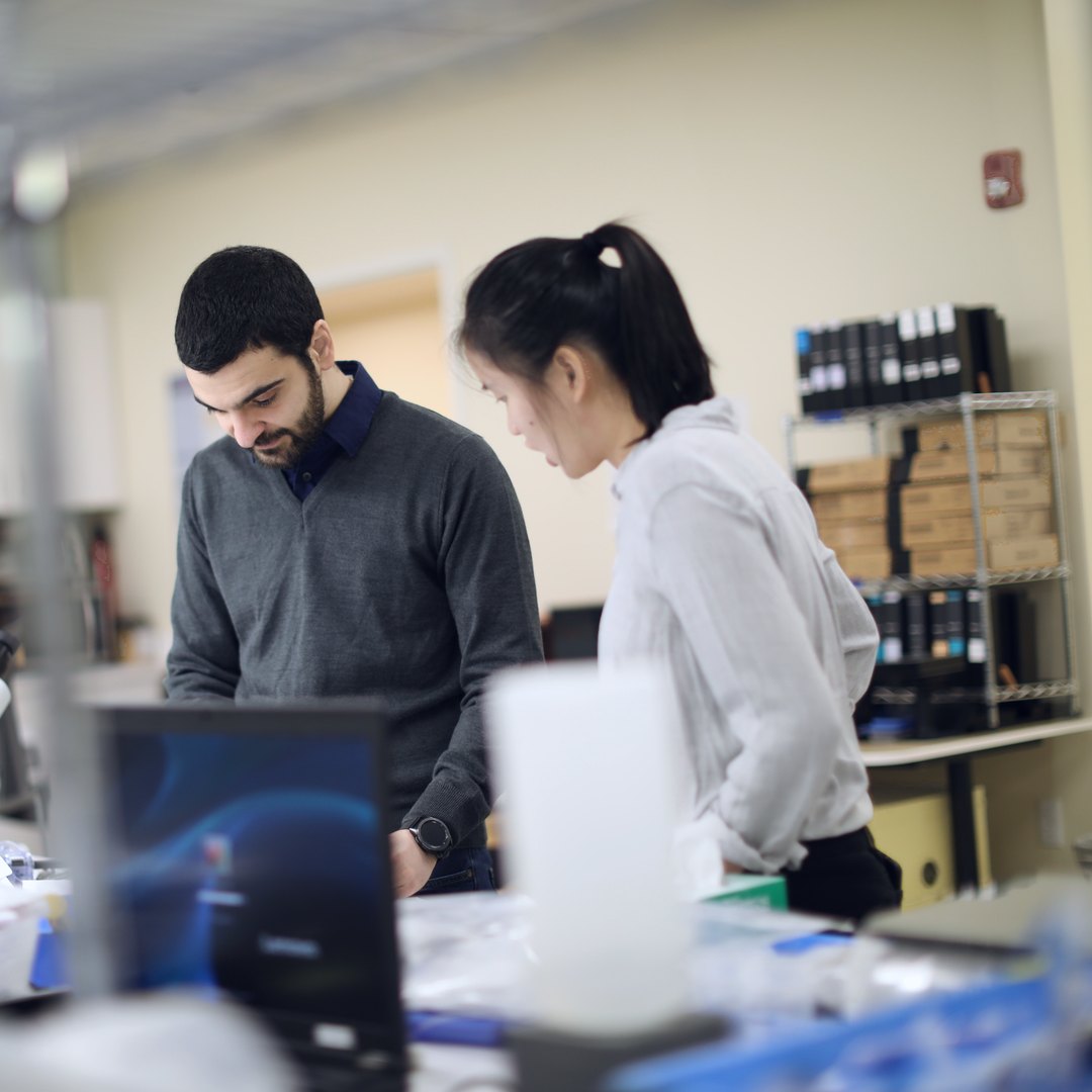 Two team members review a molded plastic component beside technical drawings on a manufacturing floor.